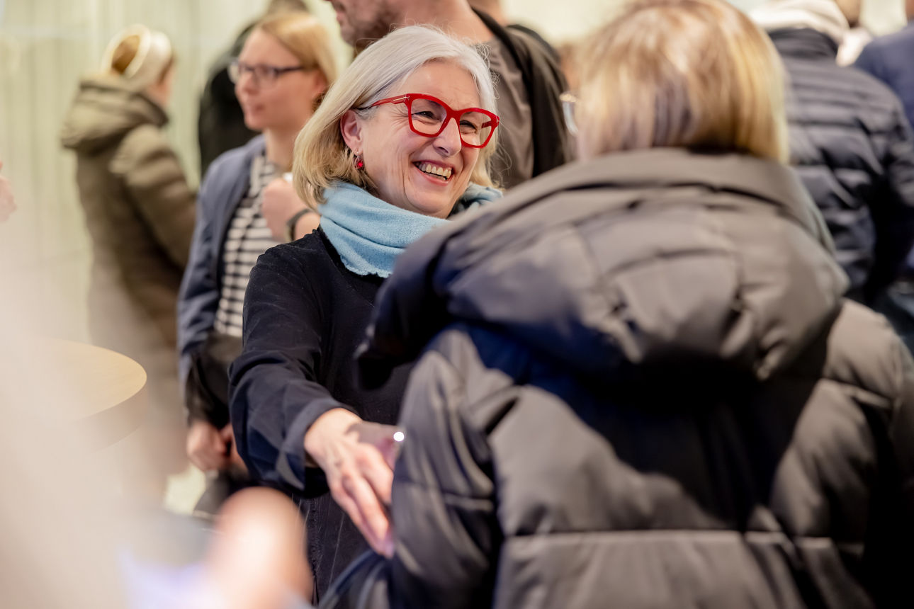 Eine lächelnde Frau mit roter Brille und hellblauem Schal unterhält sich bei einer Veranstaltung angeregt mit anderen Gästen.