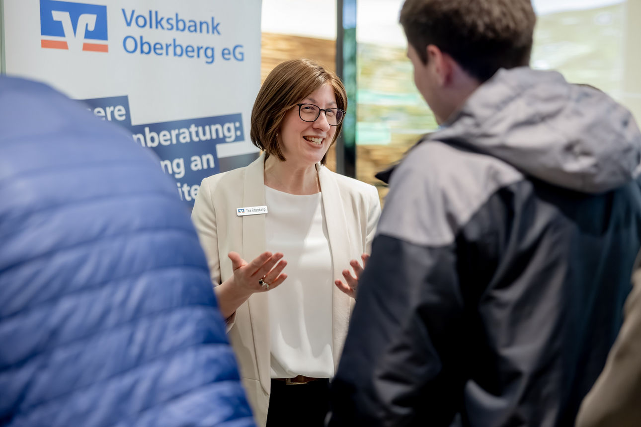 Eine Beraterin der Volksbank Oberberg eG im Gespräch mit einem jungen Mann vor einem Informationsaufsteller.