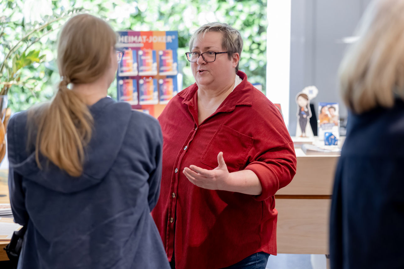 Eine Frau in einer roten Bluse spricht gestikulierend mit zwei anderen Personen bei einer Veranstaltung.