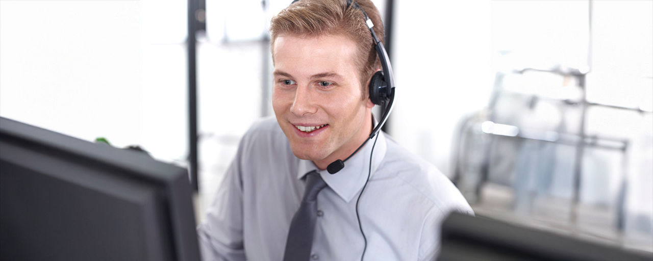 Mann sitzt lächelnd am Schreibtisch und telefoniert mit einem Headset