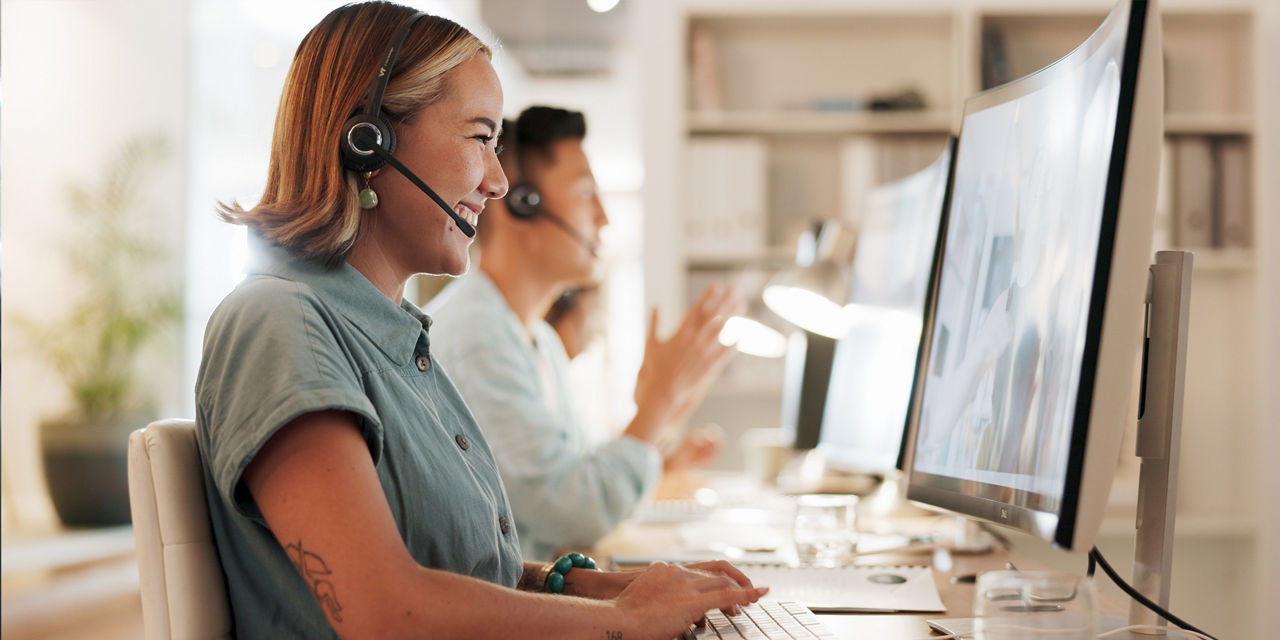 Zwei Personen mit Headsets arbeiten lächelnd an Computern in einem Büro