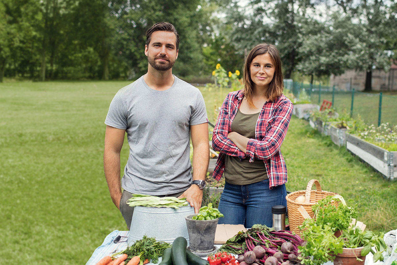 Ein Mann und eine Frau stehen in einem großen Garten, vor ihnen geerntetes Gemüse