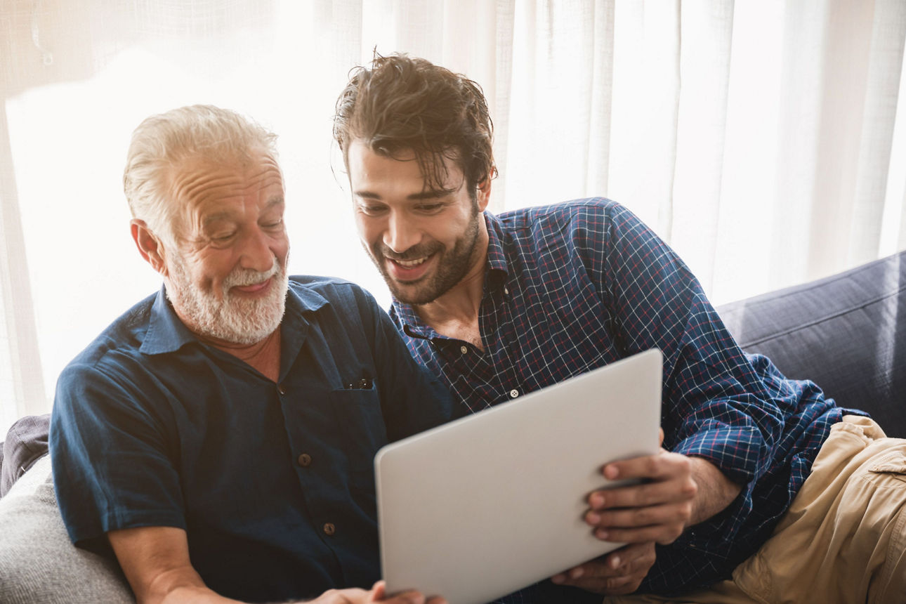Ein erwachsener Sohn und ein älterer Vater sitzen zu Hause, sehen auf ein Tablet  und unterhalten sich