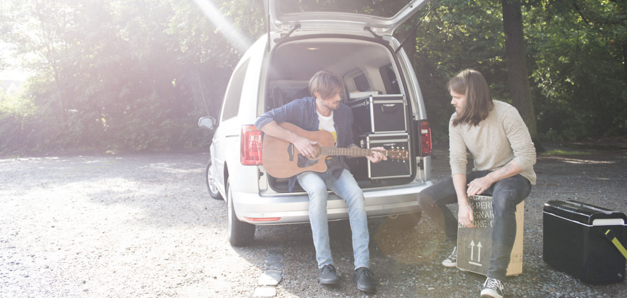 Zwei Männer mit Musikinstrumenten vorm bzw. im Kofferraum eines Autos