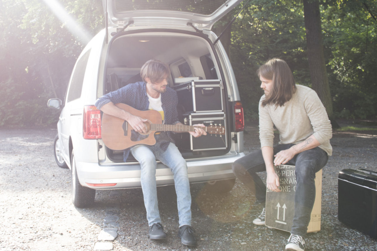 Zwei Männer mit Musikinstrumenten vorm bzw. im Kofferraum eines Autos