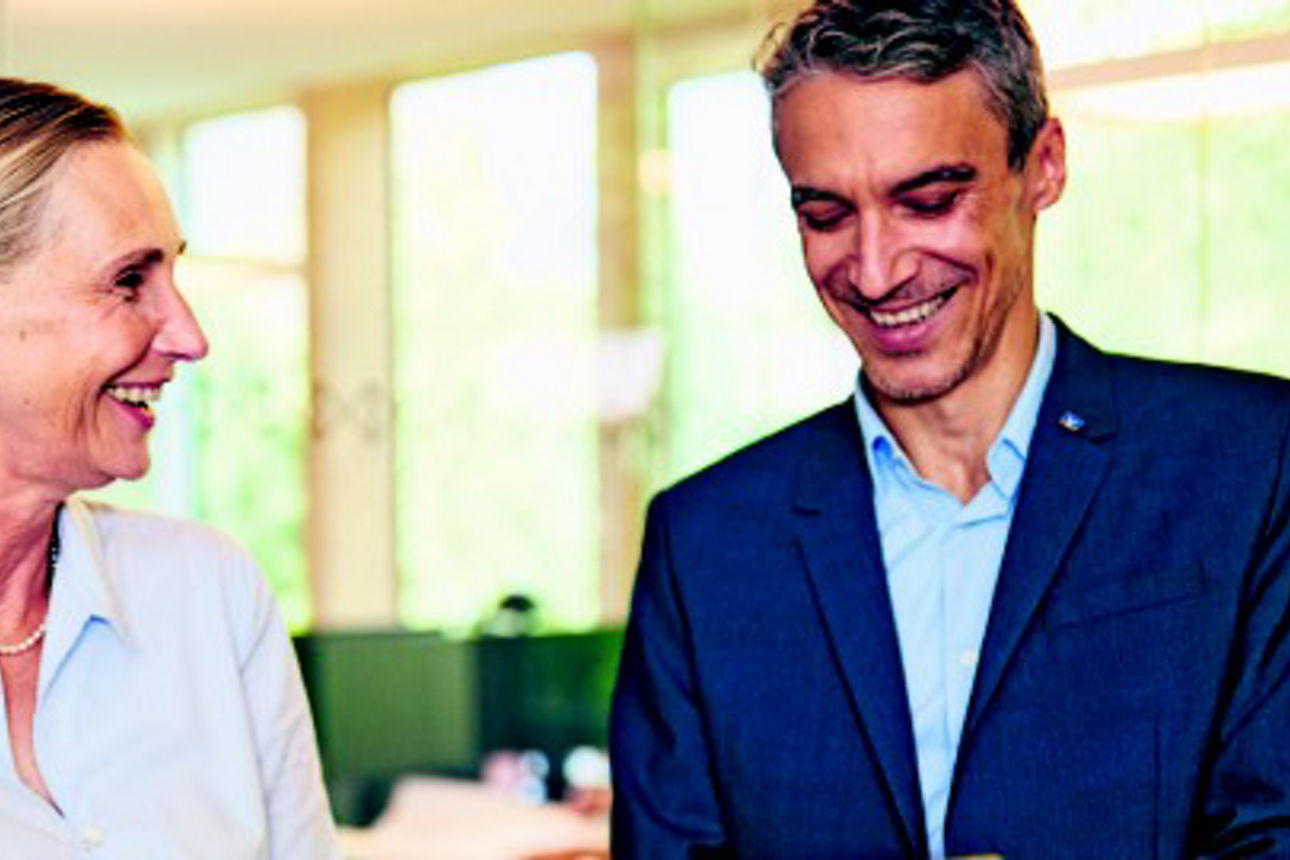 Ein Mann und eine Frau stehen in einem hell erleuchteten Büroraum und führen ein Gespräch.