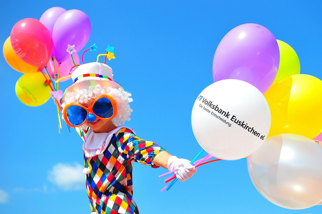 Ein als Clown verkleidetes Kind hält Luftballons in die Luft.