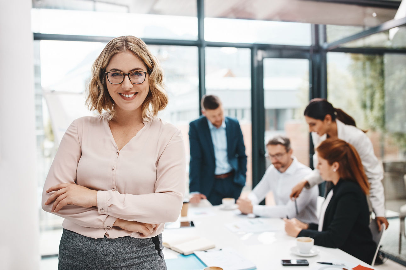 Sympathisch lächelnde Frau steht neben Konferenztisch mit anderen Kollegen dem Betrachter  zugekehrt