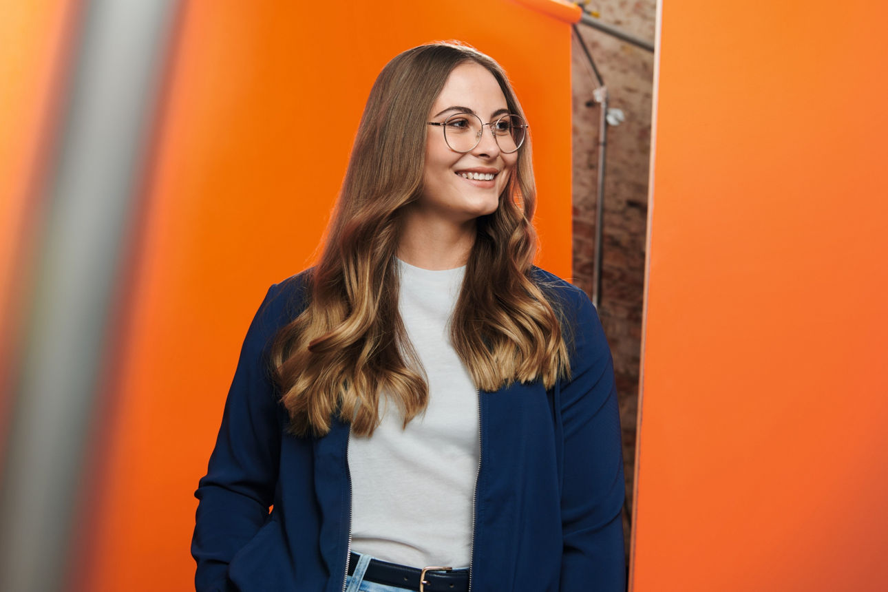 Eine junge Frau steht lächelnd vor einer organgen Wand