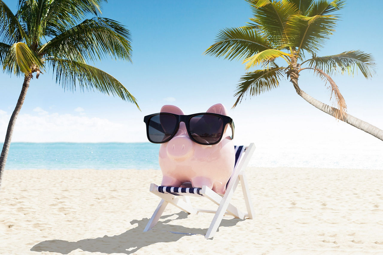 Ein rosa Sparschwein mit Sonnenbrille sitzt im Liegestuhl am Strand unter Palmen vor blauem Himmel und Meer.