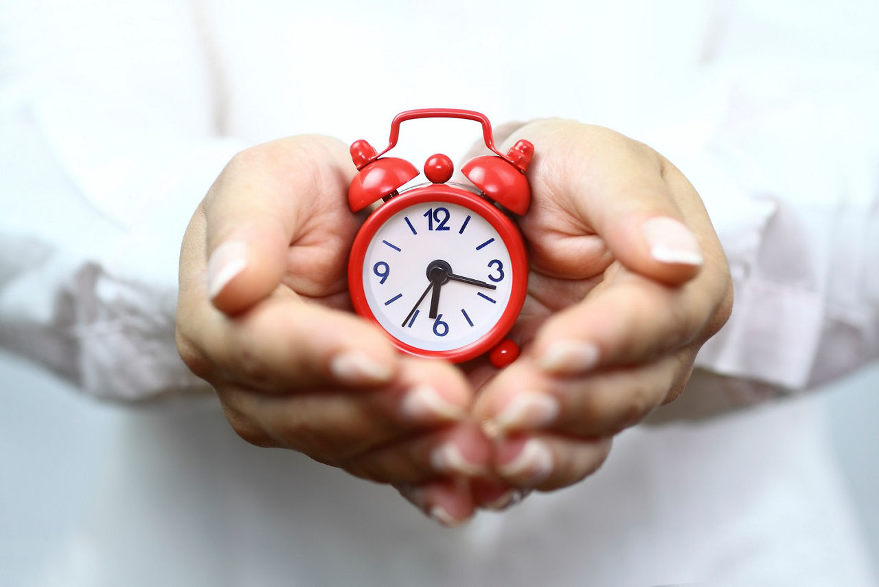 Zwei Hände halten eine kleine rote Uhr mit weißem Zifferblatt, der Hintergrund ist unscharf und hell