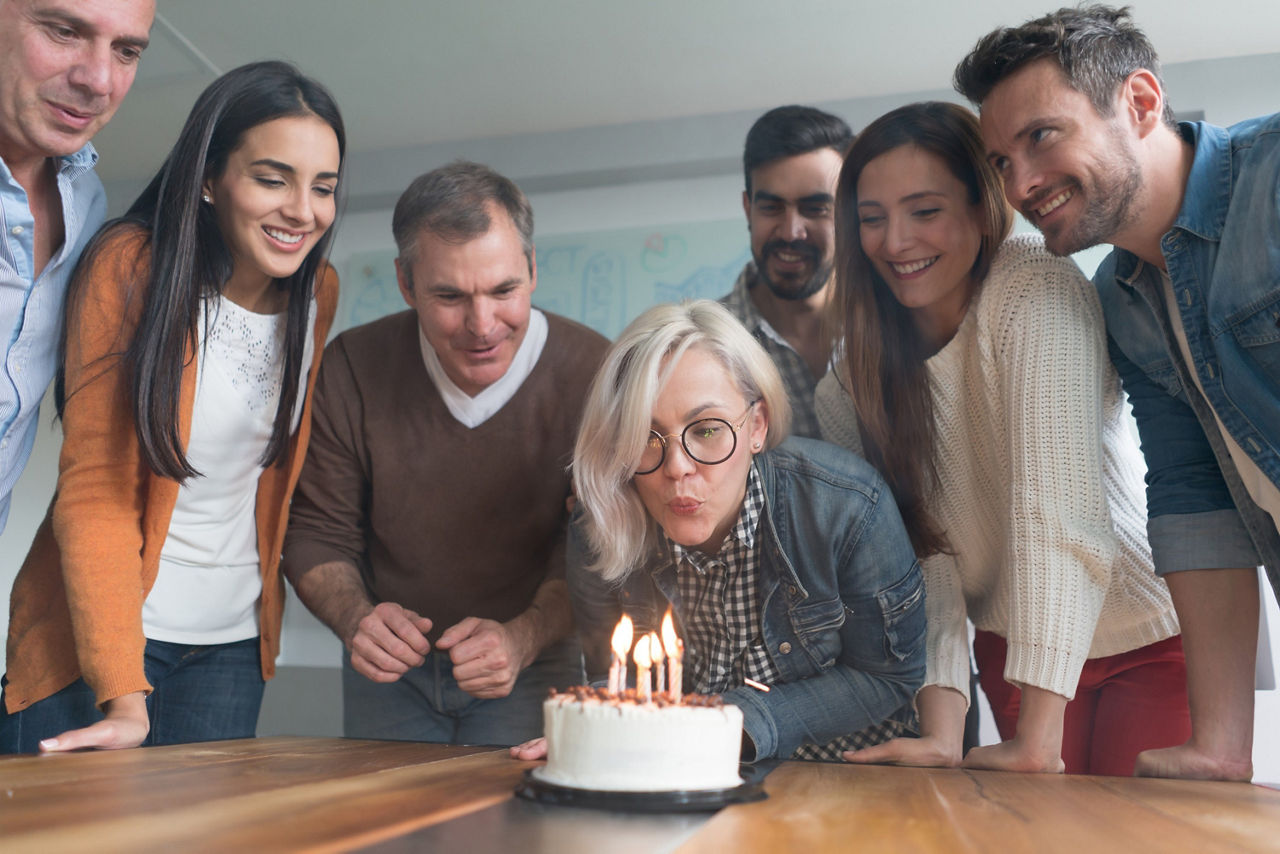Eine Gruppe personen feiert Geburtstag. Eine Frau pustet Kerzen auf dem Geburtstagskuchen aus. Die anderen Personen stehen um sie herum und lächeln.