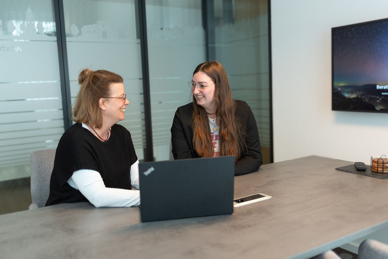 Zwei Frauen lächeln vor einem Laptop sitzend