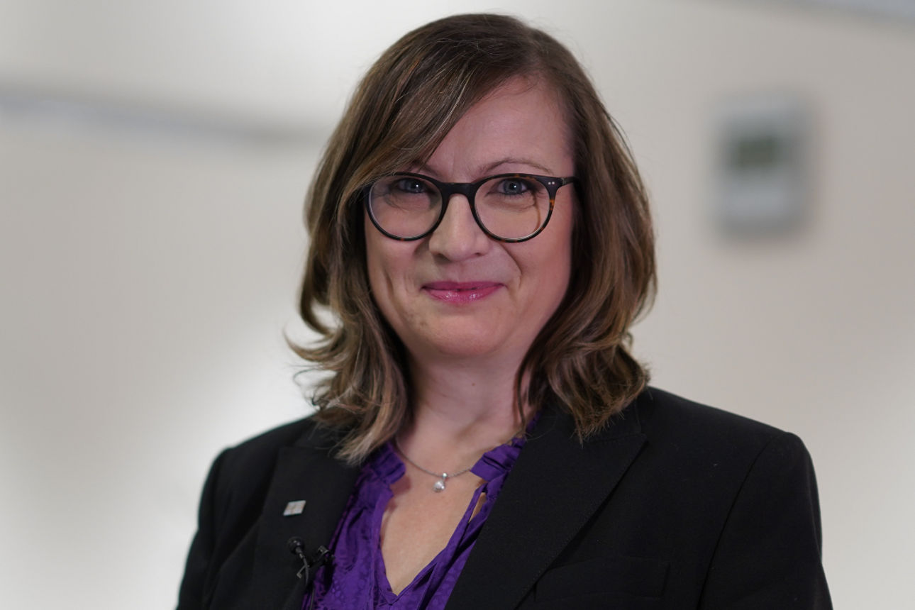 Eine Frau im Anzug steht lächelnd an ihrem Arbeitsplatz