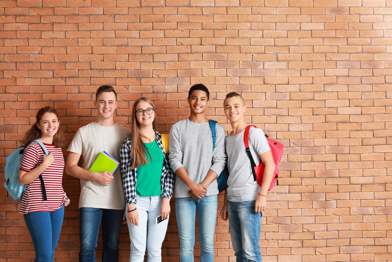 Zwei junge Schülerrinnen und drei junge Schüler stehen mit ihren Schulsachen vor einer Mauer.