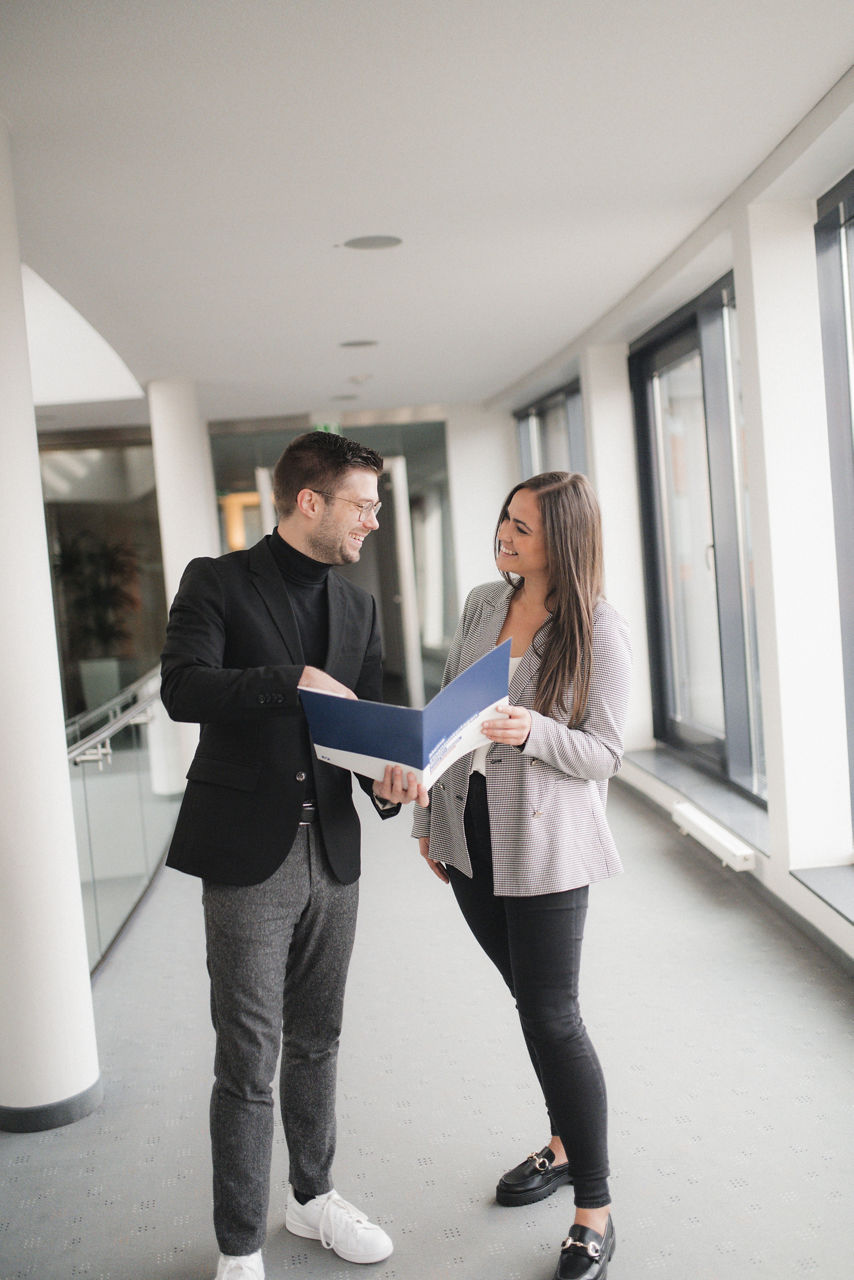 Zwei Mitarbeiter stehen auf dem Flur, halten eine Mappe in der Hand und schauen sich an.