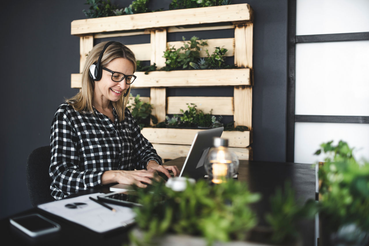 Frau mit Headset auf dem Kopf sitzt am Schreibtisch und nutzt einen Laptop