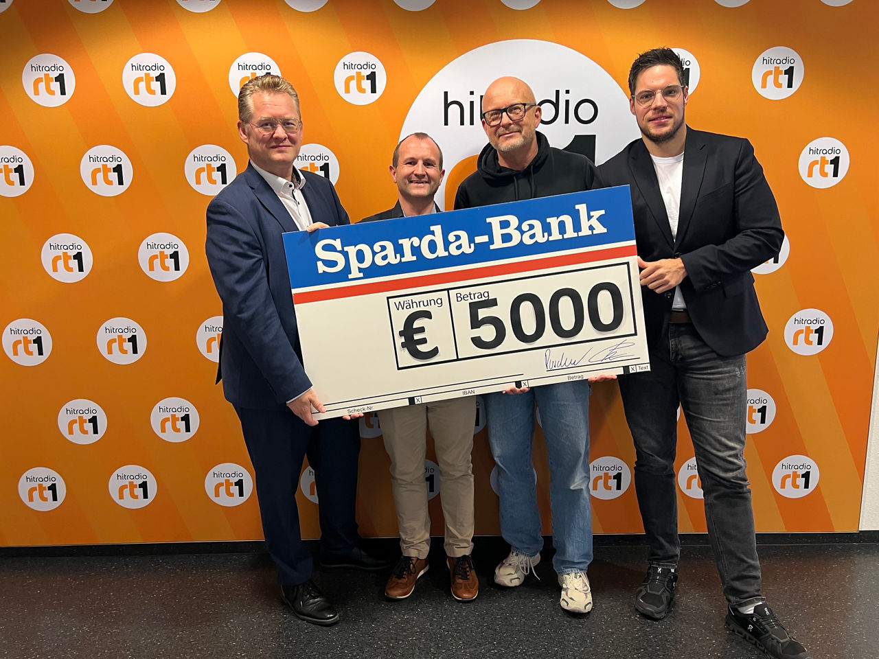 4 Personen stehen vor einer orangenen Wand und präsentieren einen großen Scheck