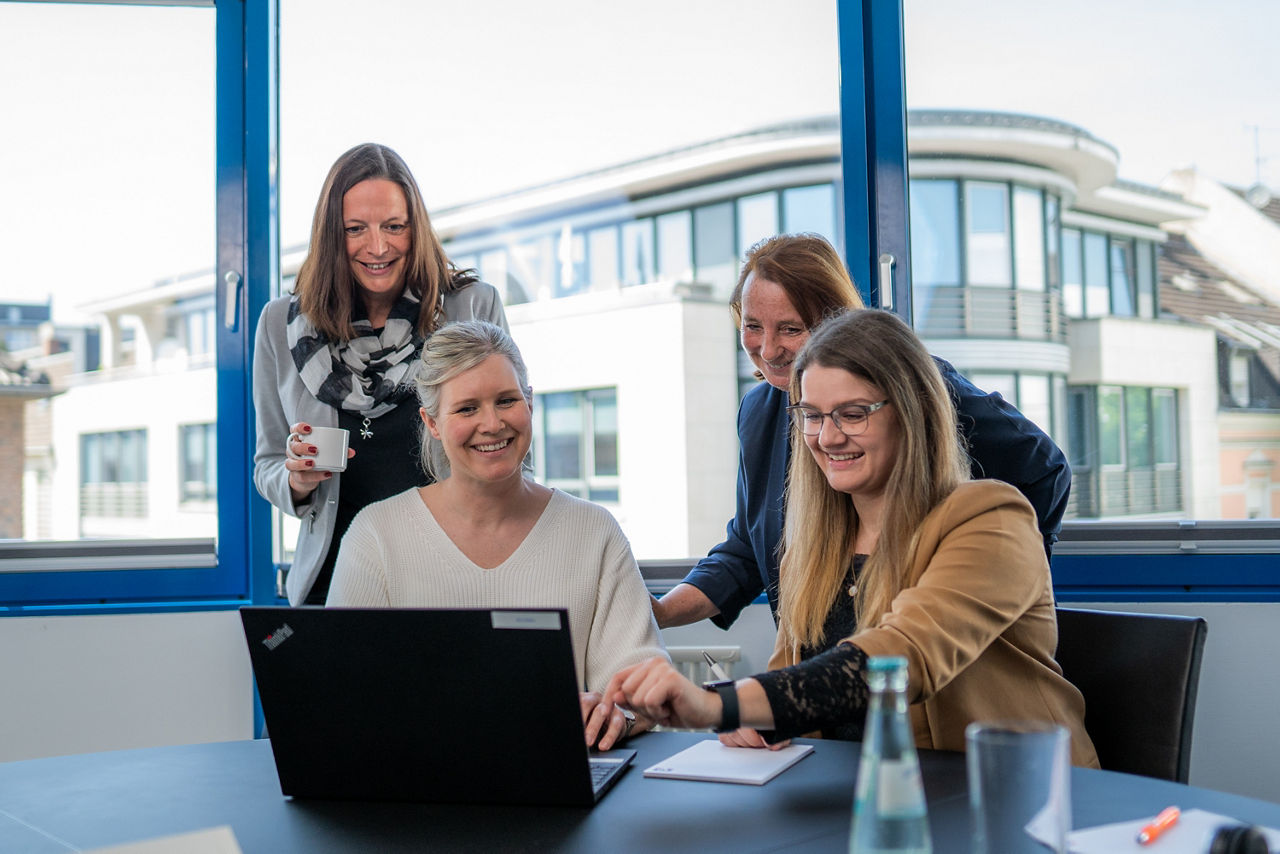 Zwei Damen sitzen in einem Büro vor einem Laptop und zwei Damen stehen dahinter