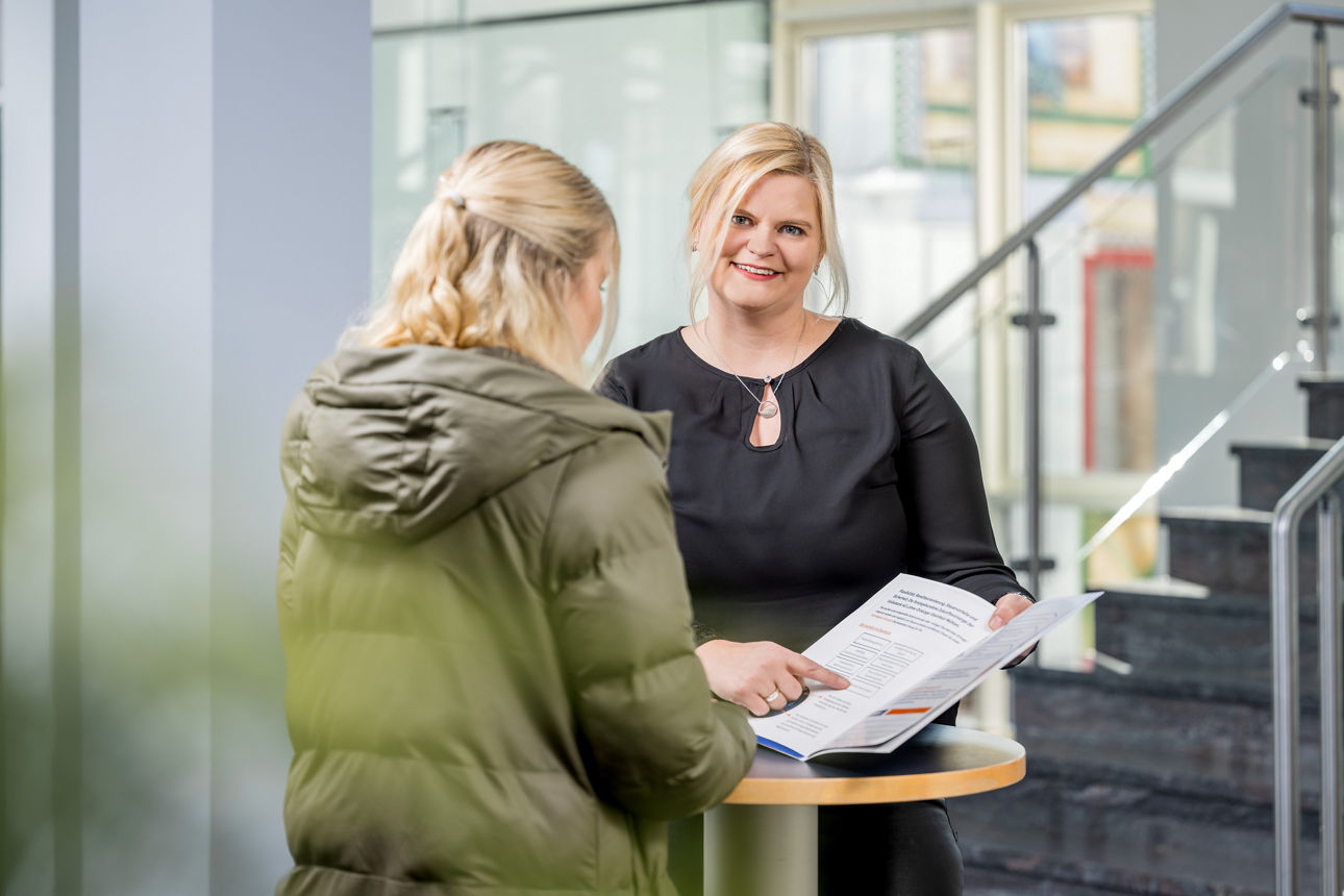 Frau steht mit anderer Frau an einem Tisch und zeigt auf eine Broschüre