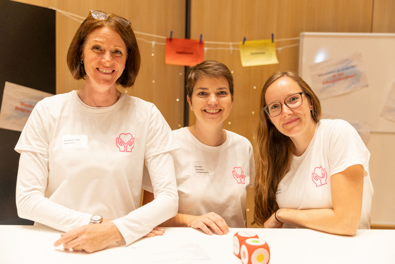 Drei Frauen stehen an einem Tisch, schauen grinsend in die Kamera. Hinter ihnen befindet sich der Aufbau eines Workshops.
