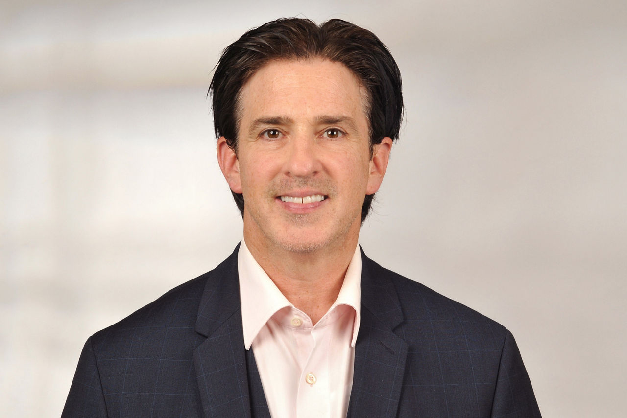 Mann mit dunklem, zurückgekämmtem Haar trägt hellrosa Hemd und dunklen Anzug vor neutralem Hintergrund