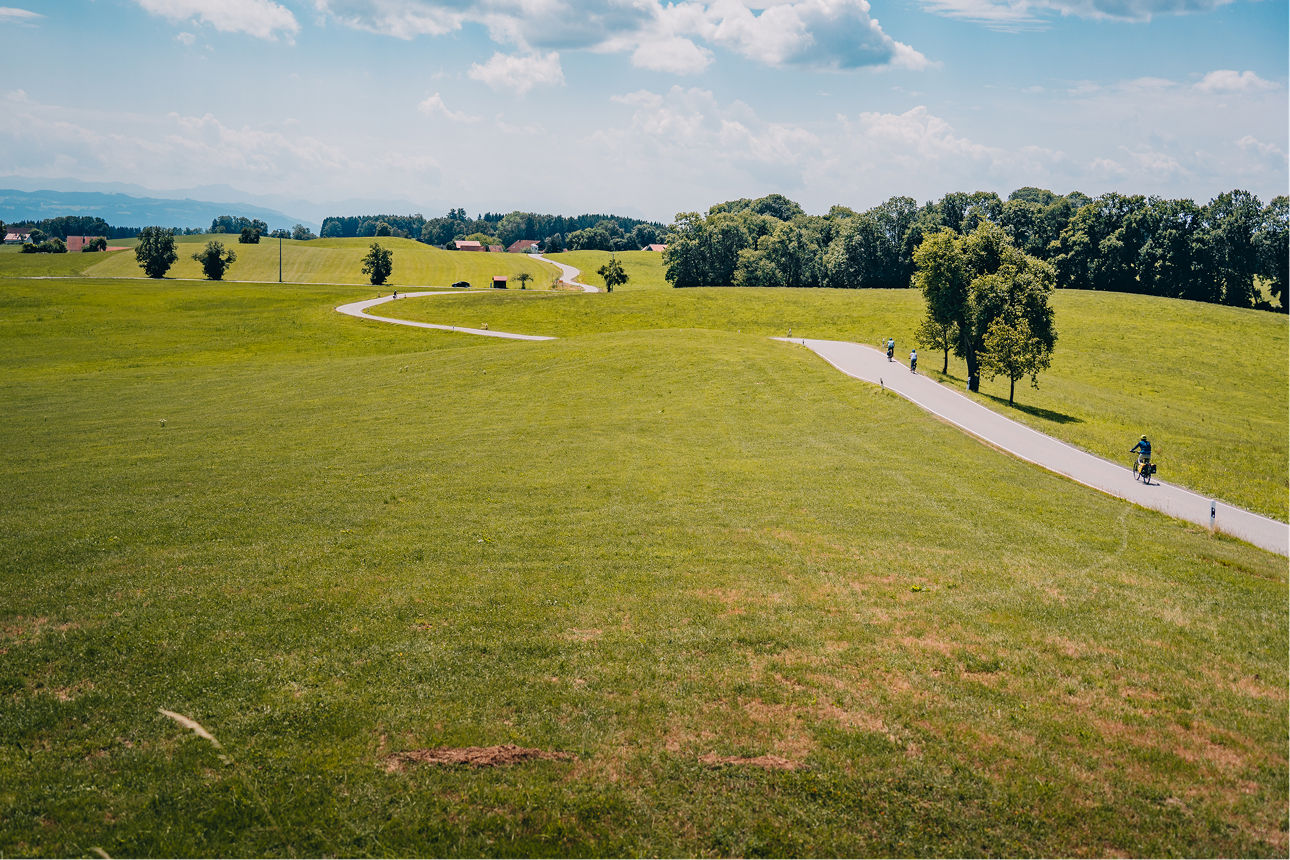 Fahrradweg, der über eine grüne hügelige Wiese verläuft, im Hintergrund sind Berge zu sehen und ein blauer wolkiger Himmel