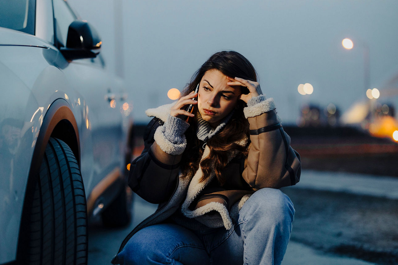 Frau sitzt vor dem Auto und telefoniert nach einer Panne.
