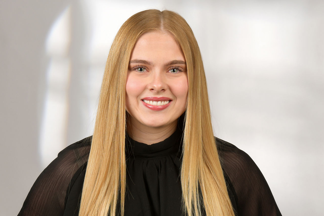 Frau mit langen, glatten blonden Haaren trägt eine schwarze Bluse mit Stehkragen vor hellem Hintergrund