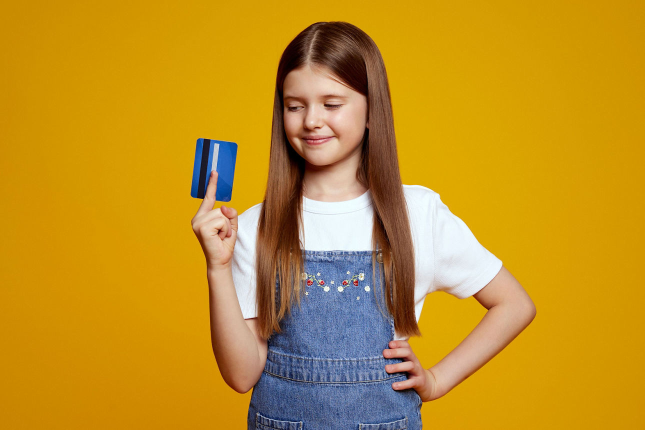 Ein Mädchen vor gelben Hintergrund schaut auf eine Girocard in ihrer rechten Hand und lächelt. 