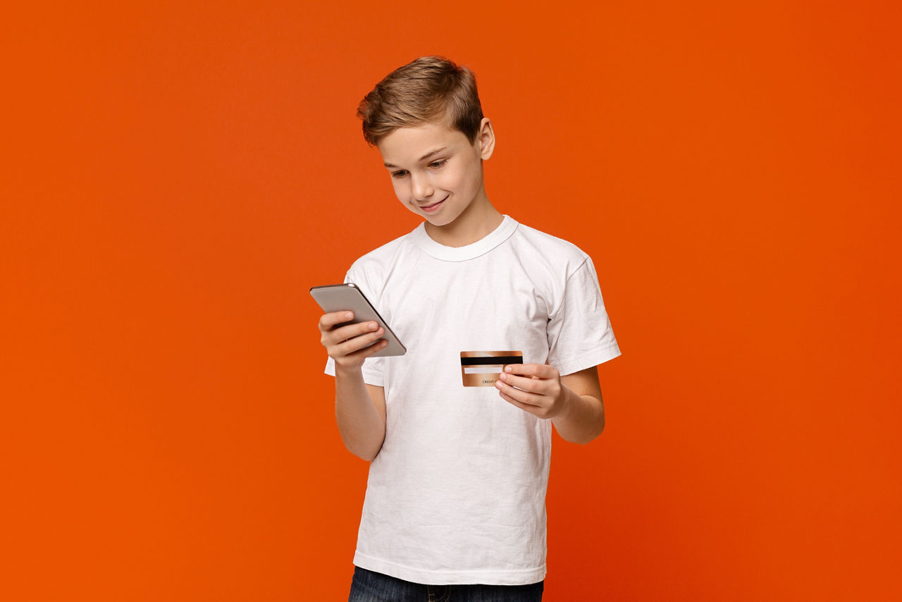 Smiling teenage boy holding credit card and smartphone in both hands, buying via internet, orange studio background