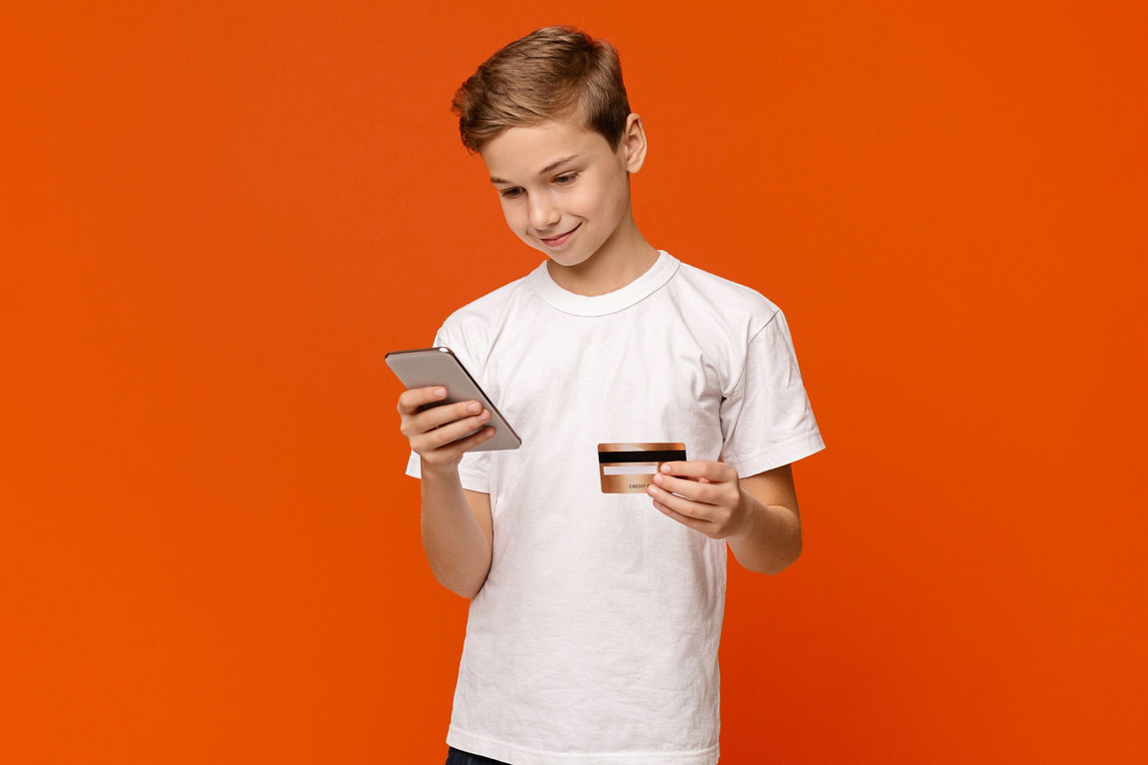 Smiling teenage boy holding credit card and smartphone in both hands, buying via internet, orange studio background