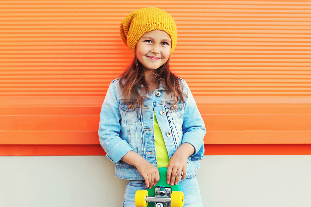 Das Bild zeigt ein lachendes Mädchen mit gelber Mütze und Skateboard vor einer orangefarbenen Wand.