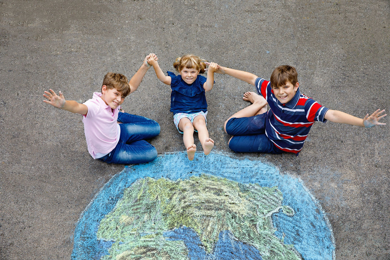 Drei Kinder sitzen Hand in Hand auf dem Boden hinter einer mit Kreide auf den Asphalt gemalten Erdkugel und winken fröhlich.