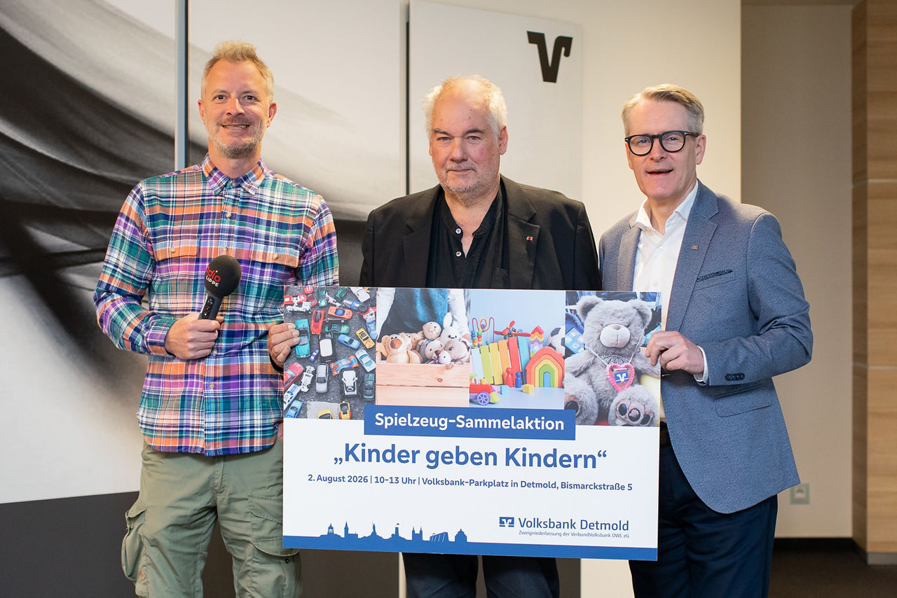 Ein Vertreter von Radio Lippe, ein Vertreter vom Verein Kinder in Not e.V. Lippe, und ein Vorstandsmitglied der VerbundVolksbank OWL eG halten ein Plakat für die Spielzeug-Sammelaktion in den Händen.