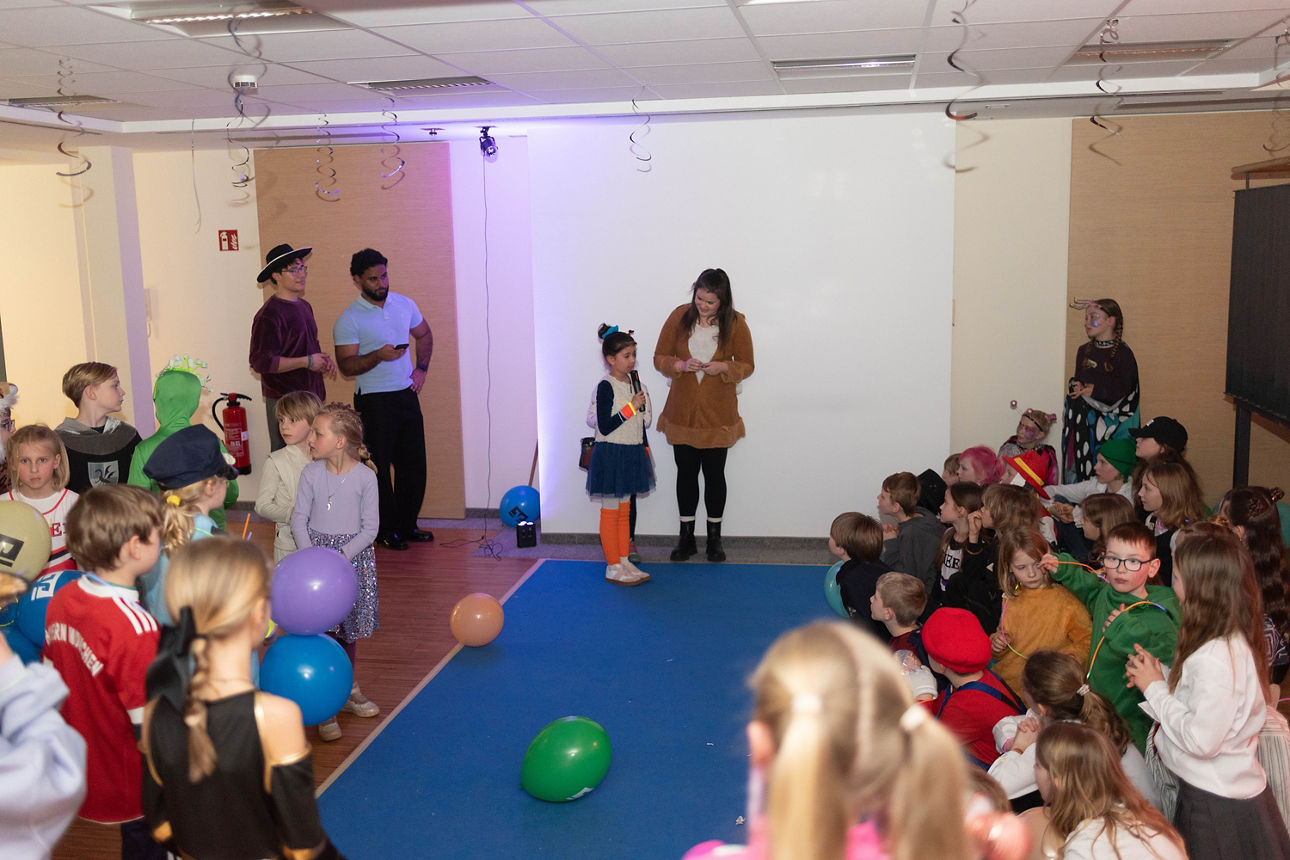 Ein verkleidetes Mädchen gibt ein Interview. Weitere verkleidete Kinder sitzen aussen herum.