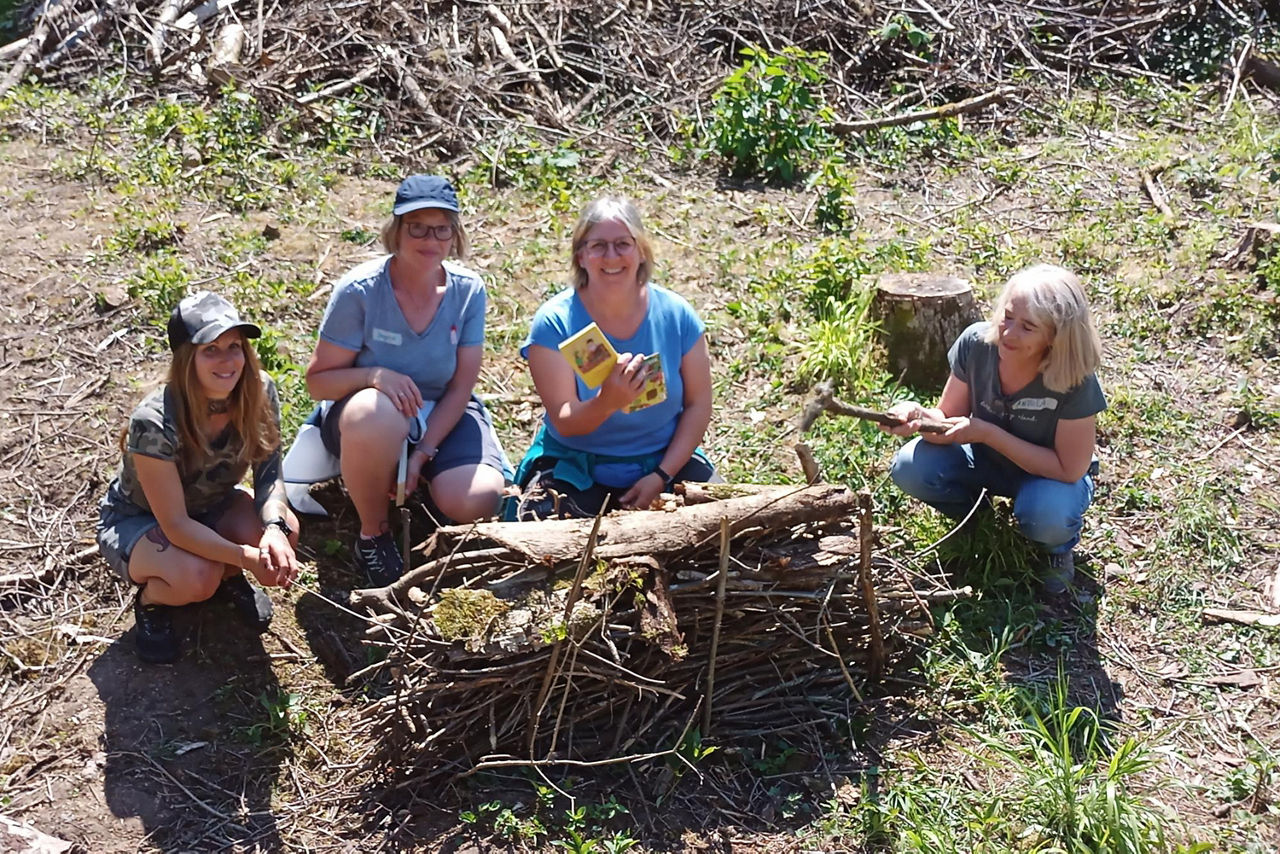Vier Frauen sitzen in einer kleinen Gruppe im Freien und bauen aus Ästen, Rinde und Moos ein Insektenhotel. Eine Teilnehmerin hält ein Naturbuch hoch und lächelt.