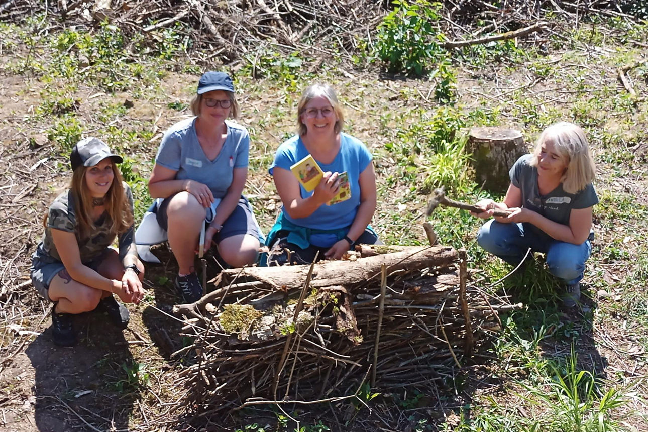 Vier Frauen sitzen in einer kleinen Gruppe im Freien und bauen aus Ästen, Rinde und Moos ein Insektenhotel. Eine Teilnehmerin hält ein Naturbuch hoch und lächelt.
