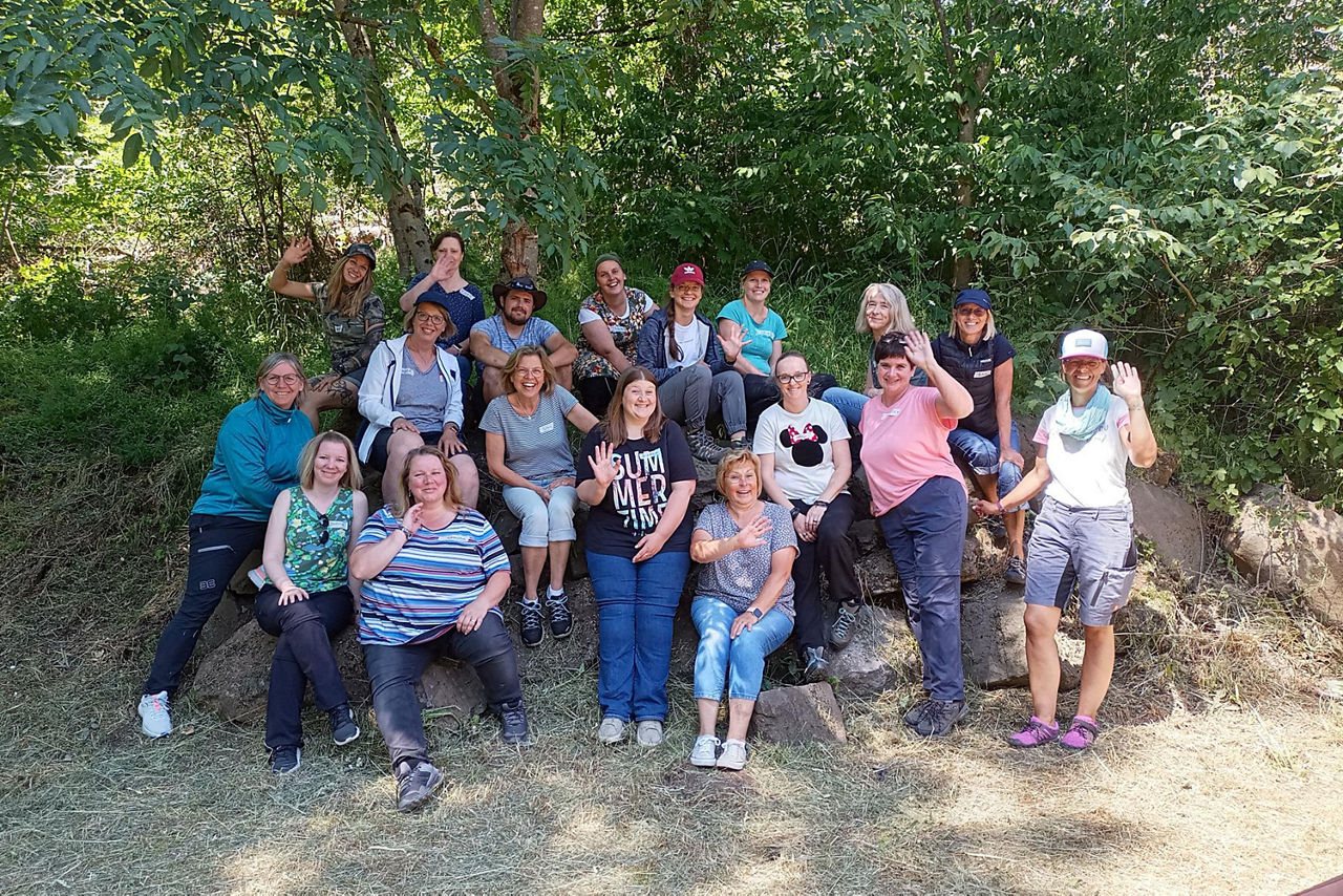 Erwachsene Teilnehmerinnen und Teilnehmer eines Workshops sitzen in einer großen Gruppe auf einer Wiese im Schatten von Bäumen. Sie lächeln und winken in die Kamera.