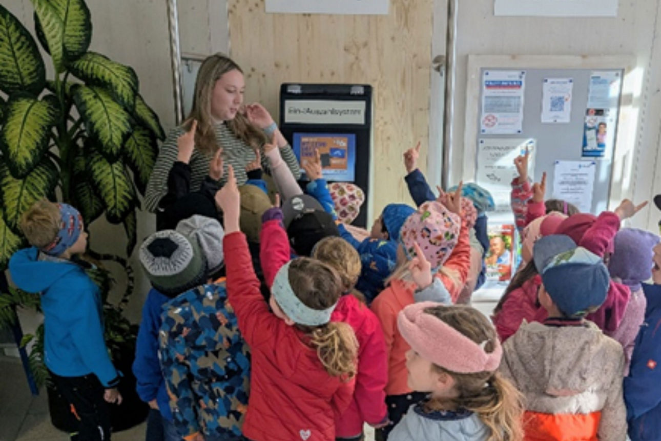 Kindergartenkinder drängen sich in einer Bankfiliale um eine erwachsene Frau und strecken ihre Finger in die Luft