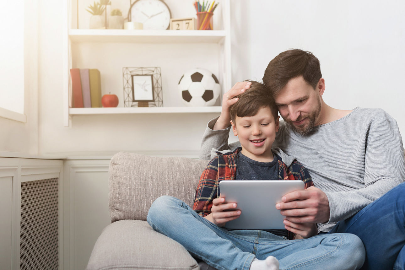 Mann zeigt seinem Sohn etwas am Tablet auf dem Sofa