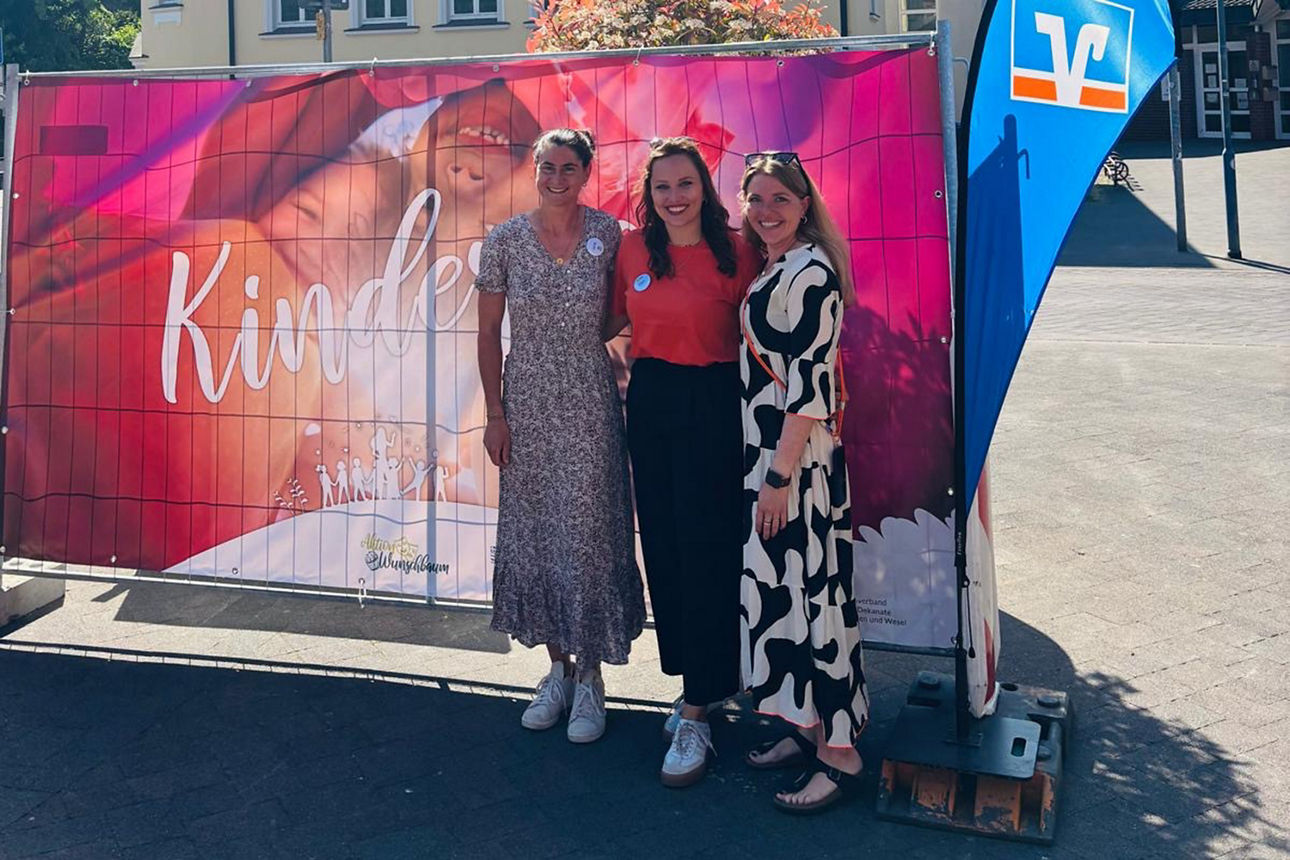 Katharina Zenker (Caritas), Ellen Weber (Gemeinde Schermbeck) und Anna Schlichting (Volksbank Schermbeck) stehen vor einem Wimpel der Volksbank Schermbeck und dem Banner zum Kindertag