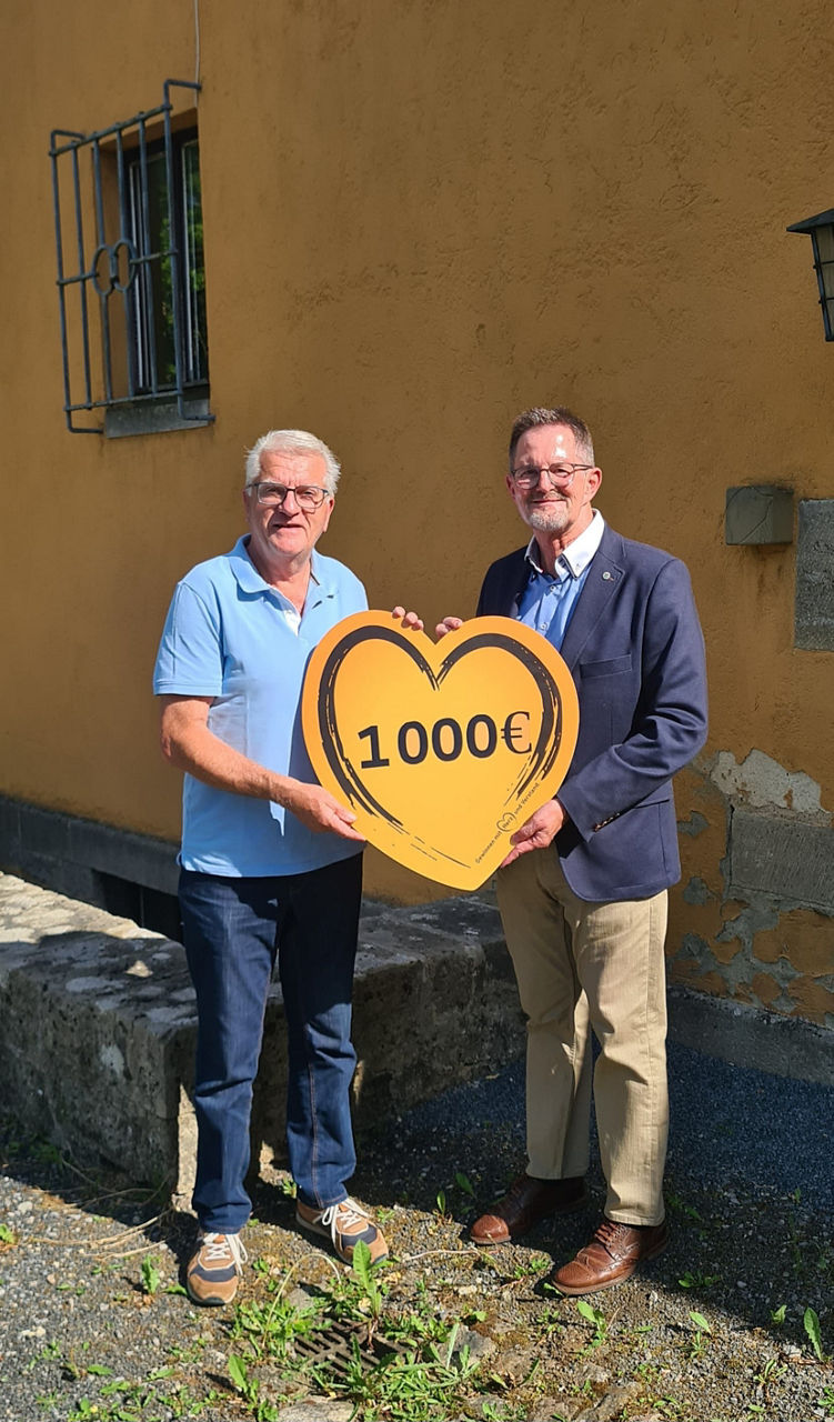 Zwei Männer stehen vor einer Kirchenmauer und halten gemeinsam ein großes Herz mit der Aufschrift „1000 €“ – eine symbolische Spendenübergabe der VR-Bank Main-Rhön an die Kirchenstiftung Unsleben.