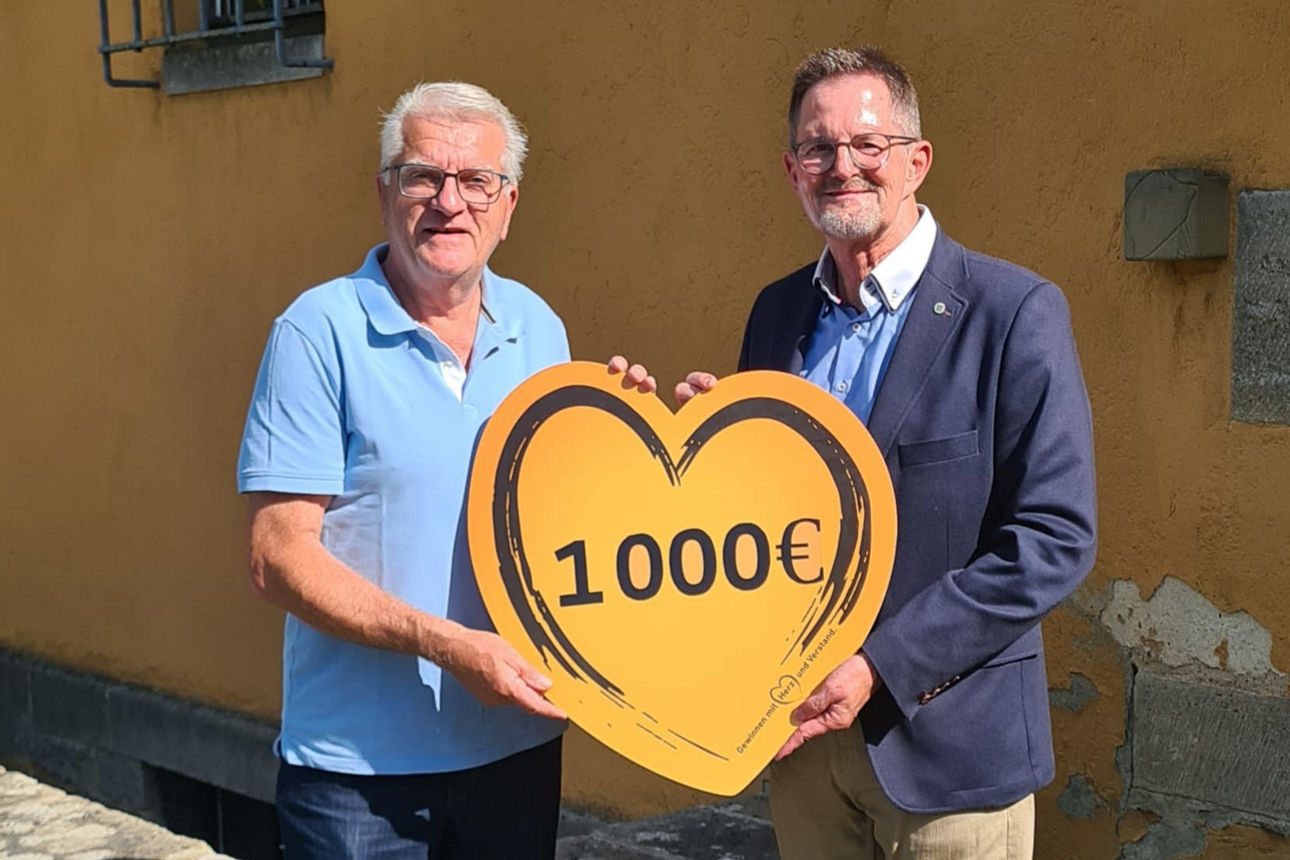Zwei Männer stehen vor einer Kirchenmauer und halten gemeinsam ein großes Herz mit der Aufschrift „1000 €“ – eine symbolische Spendenübergabe der VR-Bank Main-Rhön an die Kirchenstiftung Unsleben.