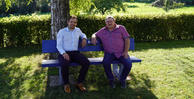 Zwei Männer sitzen auf einer blauen Bank auf einer Wiese.