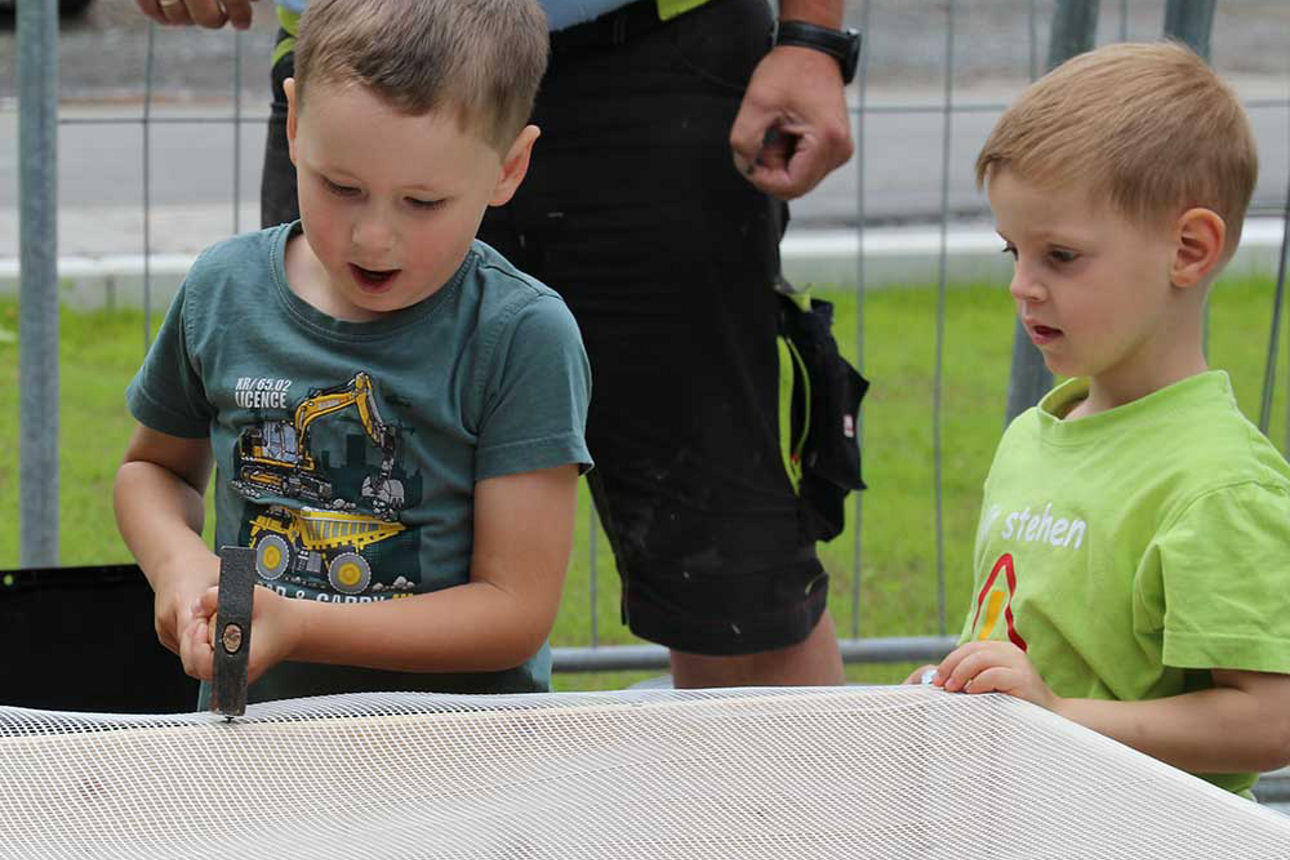 Ein Junge befestigt einen Stoff an einem Hochbeet. Er schlägt mit einem Hammer einen Nagel in das Holz.