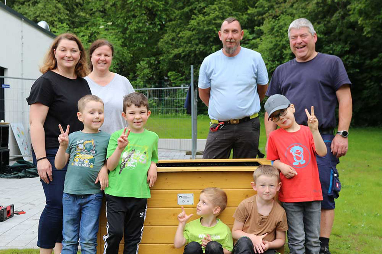 Das Bild zeigt fünf Kinder und vier Erwachsene, die um ein Hochbeet herumstehen und sich freuen.