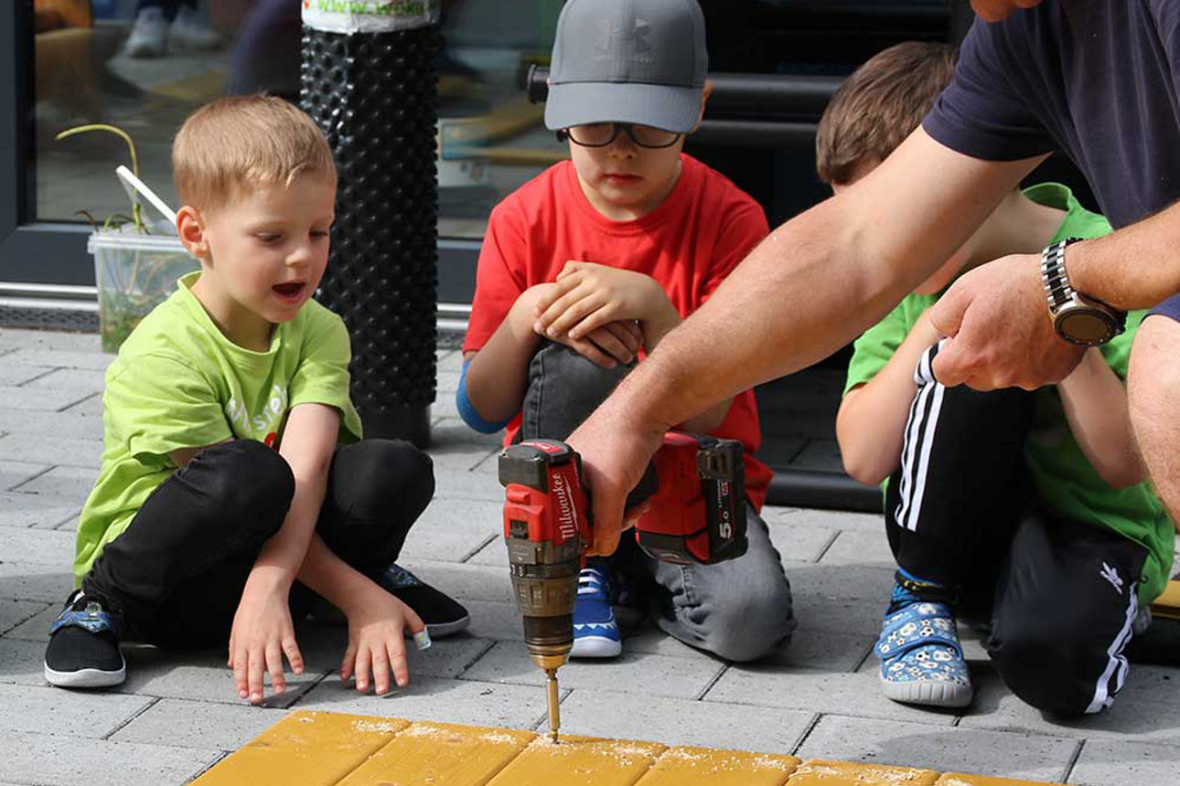 Auf dem Boden liegen Bretter. Ein Mann befestigt mit einem Akku-Schrauber Schrauben, während ihm zwei Kinder dabei zusehen.