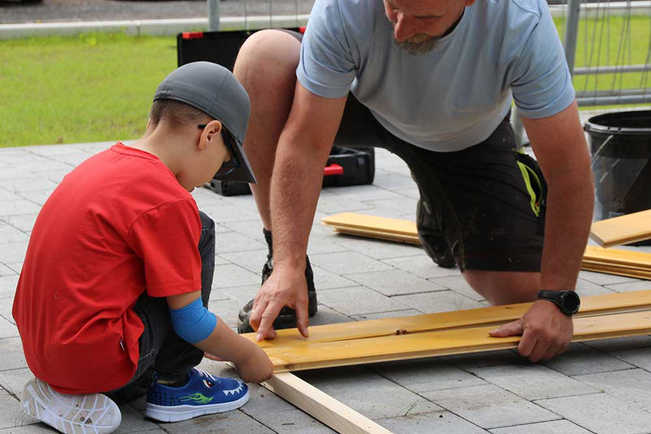 Das Bild zeigt einen Jungen und einen Mann. Sie knien auf dem Boden über den Brettern des Hochbeets.