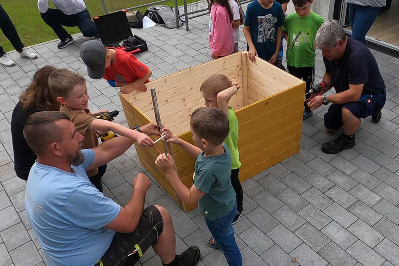 Ein unbefülltes Hochbeet steht auf dem Boden. Ein Mann hält einen Zollstock an, während im drei Kinder zusehen.